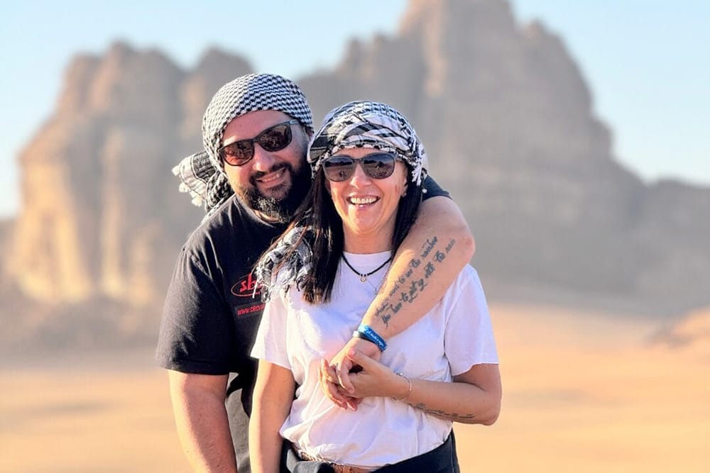 Un uomo e una donna sorridenti con occhiali da sole e foulard a scacchi posano insieme in un soleggiato paesaggio desertico con formazioni rocciose, catturando un momento di gioia durante il Viaggio Giordania Tour 2023. L'uomo ha un braccio intorno alle spalle della donna.