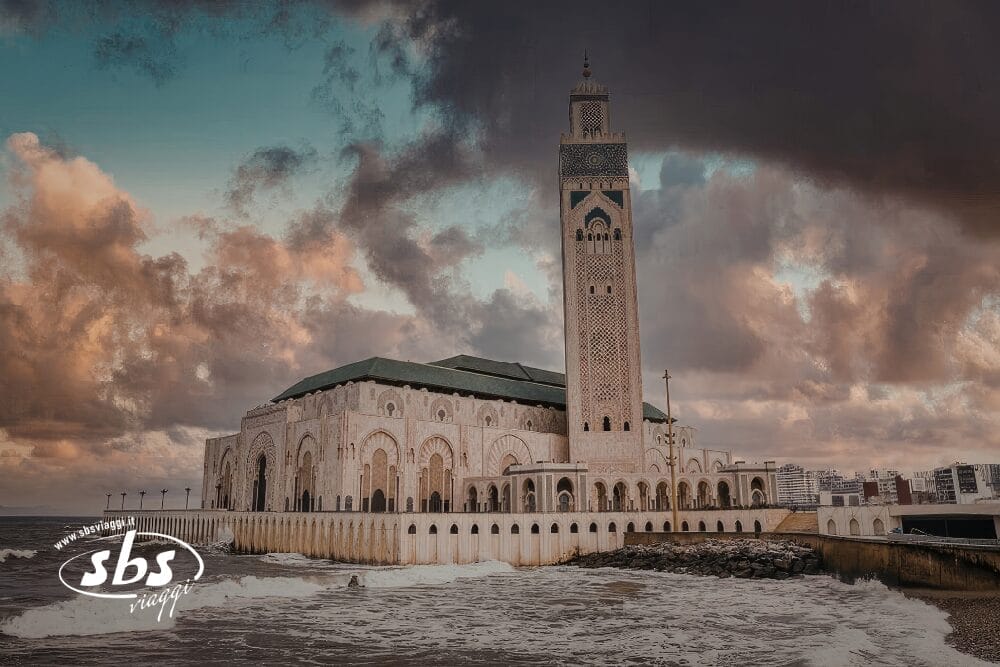 L'immagine mostra la Moschea di Hassan II a Casablanca, in Marocco, vicino all'Oceano Atlantico, con le nuvole e le onde che si infrangono contro la parete del mare, un vero Tesori del Marocco con il suo alto minareto che spicca.