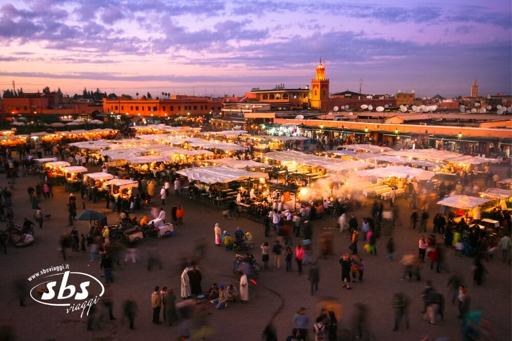 Un vivace mercato all'aperto al tramonto, pieno di gente, bancarelle illuminate e tende colorate: una scena da Best of Morocco. Sullo sfondo, la torre di una moschea si staglia sotto un cielo viola e arancione.