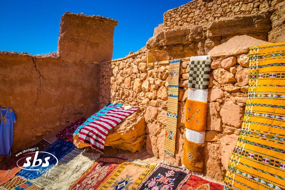 Tappeti tradizionali colorati sono appesi e posati su muri di pietra grezza e sul terreno in un mercato all'aperto illuminato dal sole, sotto un cielo azzurro e limpido. Scoprite il fascino rustico del Marocco con Tesori del Marocco in un indimenticabile tour privato.