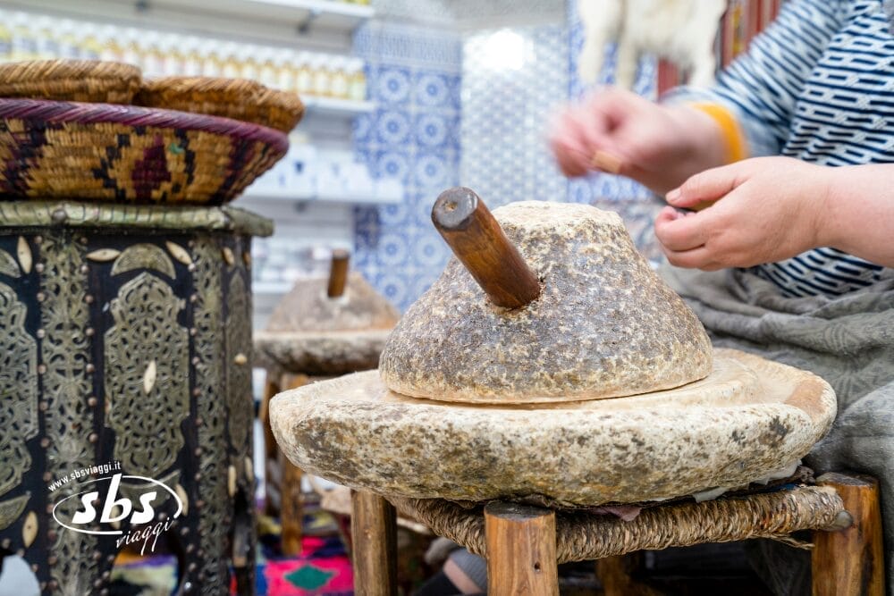 Una persona utilizza un tradizionale mulino a mano in pietra per macinare il cibo, con cesti e piastrelle decorate sullo sfondo. Sono visibili solo le mani e parte del braccio: uno spaccato della vita quotidiana in un tour in Marocco. Nell'angolo compare il logo di SBS Viaggi.