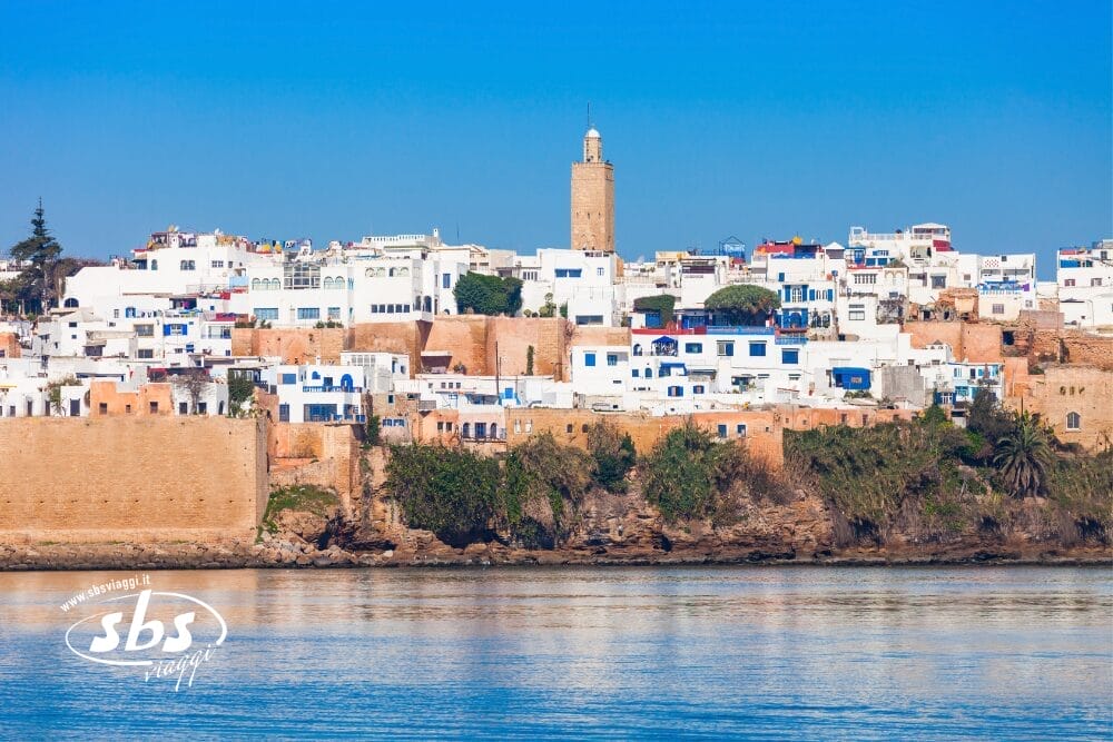 Una vista panoramica di edifici bianchi e blu a Casablanca, con un alto minareto che si erge sopra le mura storiche della città, il tutto affacciato su acque calme sotto un cielo azzurro e limpido: l'essenza di un tour Best of Morocco.