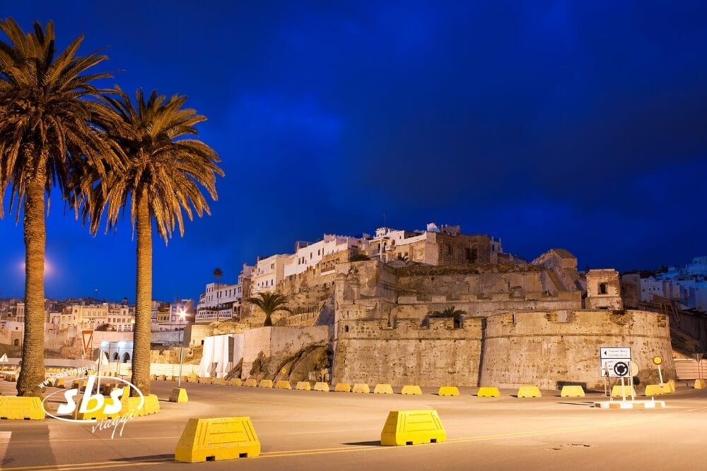 Una storica fortezza in pietra con edifici bianchi su una collina è illuminata di notte, incorniciata da palme e barriere stradali gialle sotto un cielo blu intenso: una scena straordinaria da vivere durante un tour di Casablanca, davvero il meglio del Marocco.