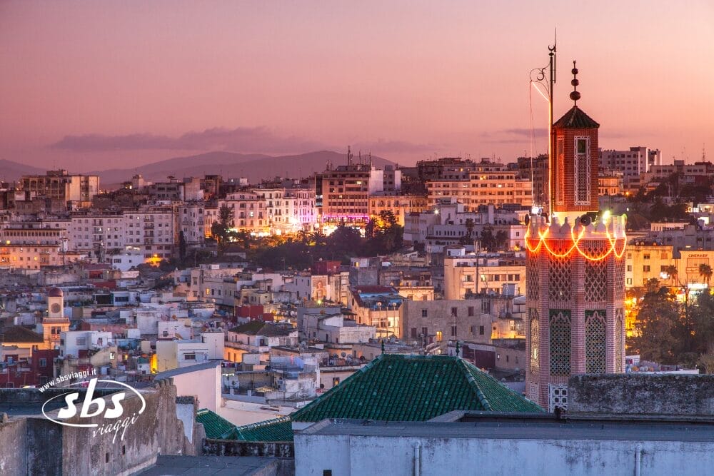 Un paesaggio urbano al tramonto con un minareto decorato di luci in primo piano, circondato da edifici bianchi e dalle luci della città in lontananza: uno scenario perfetto per scoprire i Tesori del Marocco in un indimenticabile Tour Privato Marocco, con le montagne sullo sfondo.
