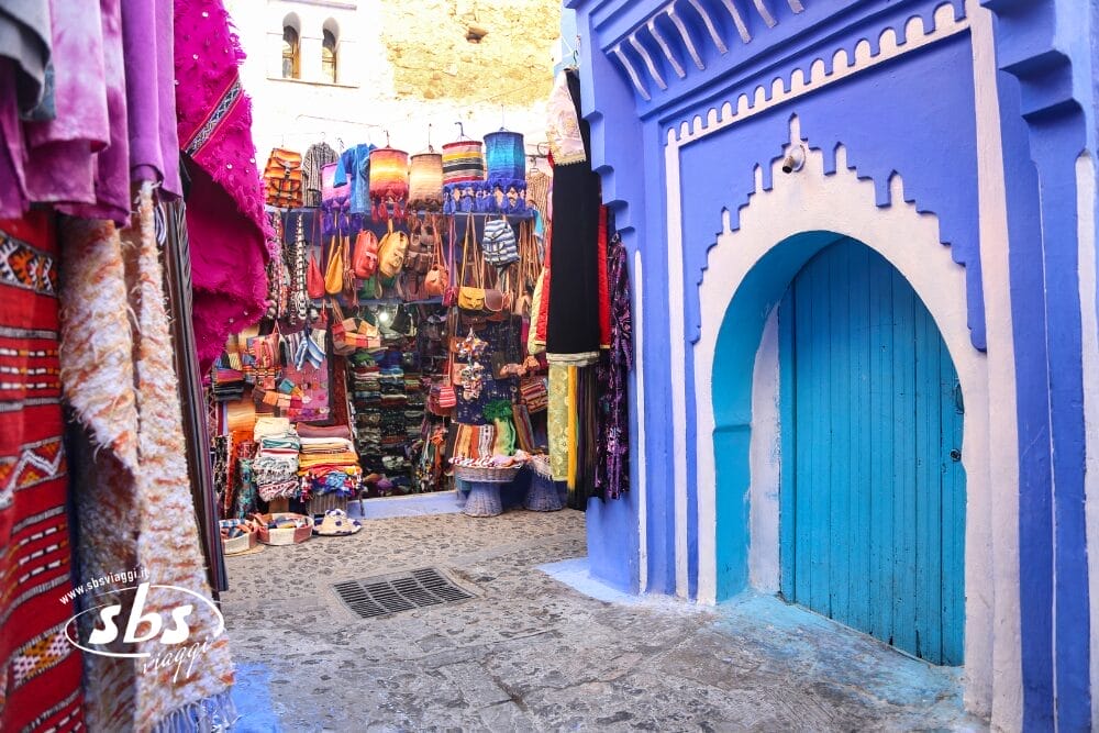 Un vivace mercato di strada, incluso nel tour Best of Morocco, con tessuti colorati, borse e cappelli esposti accanto a una porta ad arco blu in uno stretto vicolo lastricato in pietra; l'architettura tradizionale nordafricana aggiunge un fascino autentico.
