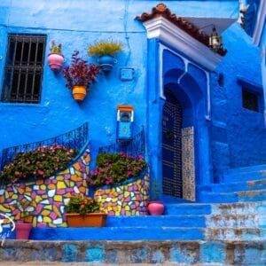 Un edificio blu vibrante a Chefchaouen, in Marocco, adornato con piastrelle a mosaico colorate e vasi di fiori vivaci. Esplorate questi Tesori del Marocco: piastrelle decorate, pareti blu vivo e ringhiere in ferro battuto vi aspettano nel vostro Tour Privato.