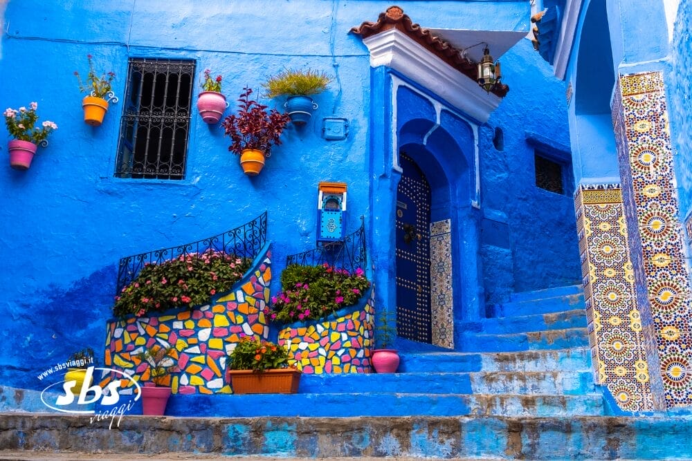 Un edificio blu vibrante a Chefchaouen, in Marocco, adornato con piastrelle a mosaico colorate e vasi di fiori vivaci. Esplorate questi Tesori del Marocco: piastrelle decorate, pareti blu vivo e ringhiere in ferro battuto vi aspettano nel vostro Tour Privato.