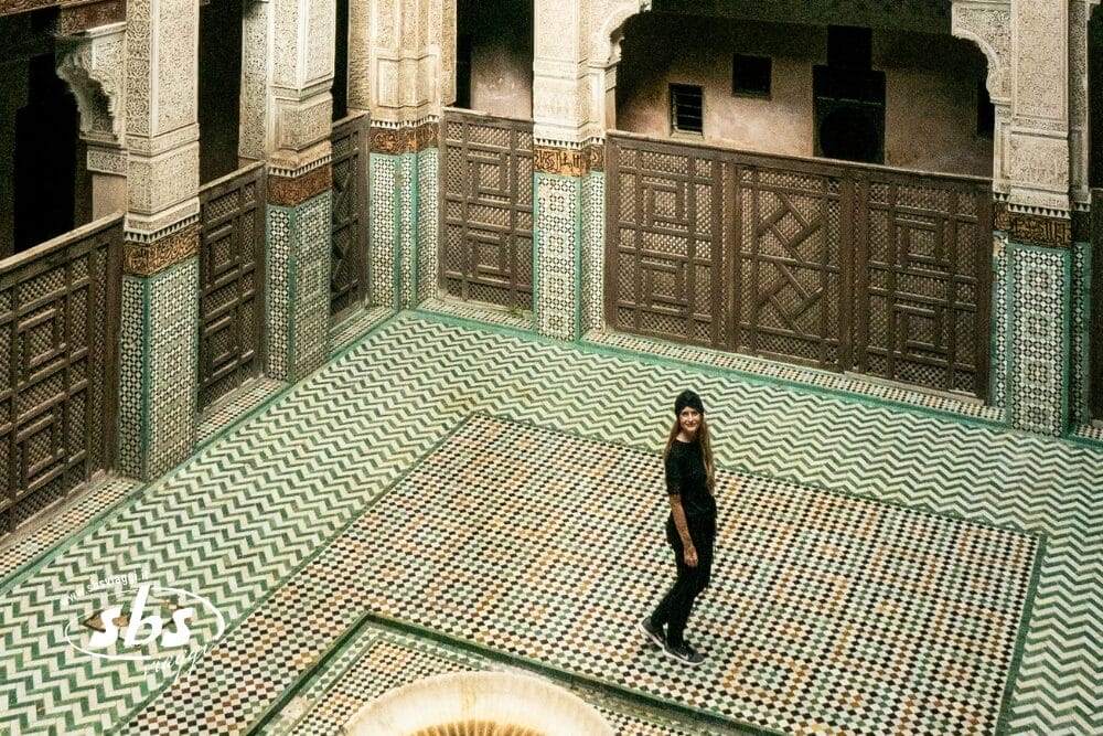 Una persona si trova in un grande cortile decorato con pavimenti in piastrelle e intricate pareti in legno e piastrelle, accanto a una piccola fontana: una scena straordinaria di un tour Best of Morocco. L'architettura tradizionale e storica cattura l'essenza del design marocchino.