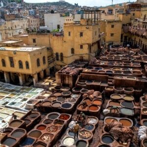 Veduta di una conceria tradizionale con molte vasche di tintura rotonde in un cortile aperto, circondato da edifici gialli. Si vedono gli operai che si occupano dei tini, con il paesaggio urbano del Marocco sullo sfondo: una vera Tesori del Marocco.