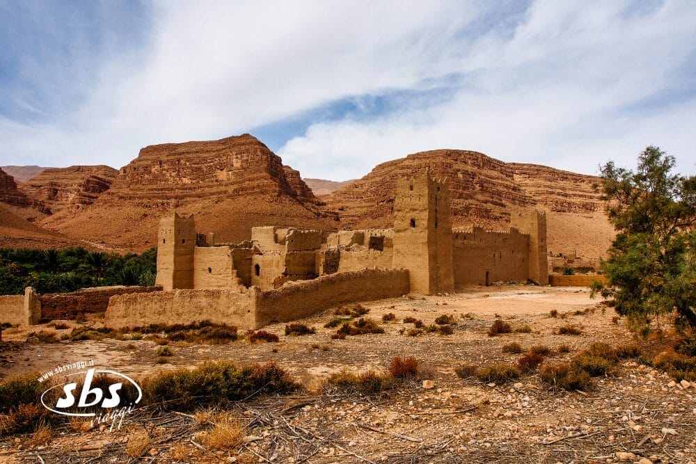 L'antica fortezza in mattoni di fango con torri multiple si erge in un paesaggio desertico arido e roccioso in Marocco, con colline aspre e vegetazione rada sullo sfondo sotto un cielo parzialmente nuvoloso: un vero punto di forza per qualsiasi tour privato di Tesori del Marocco.