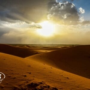 Il sole dorato tramonta su ampie dune di sabbia in Marocco, sotto un cielo drammatico. Ombre e luci evidenziano le curve del deserto, evocando la magia del Tour Privato "Tesori del Marocco". Il logo bianco "sbs viaggi" si trova nell'angolo in basso a sinistra.