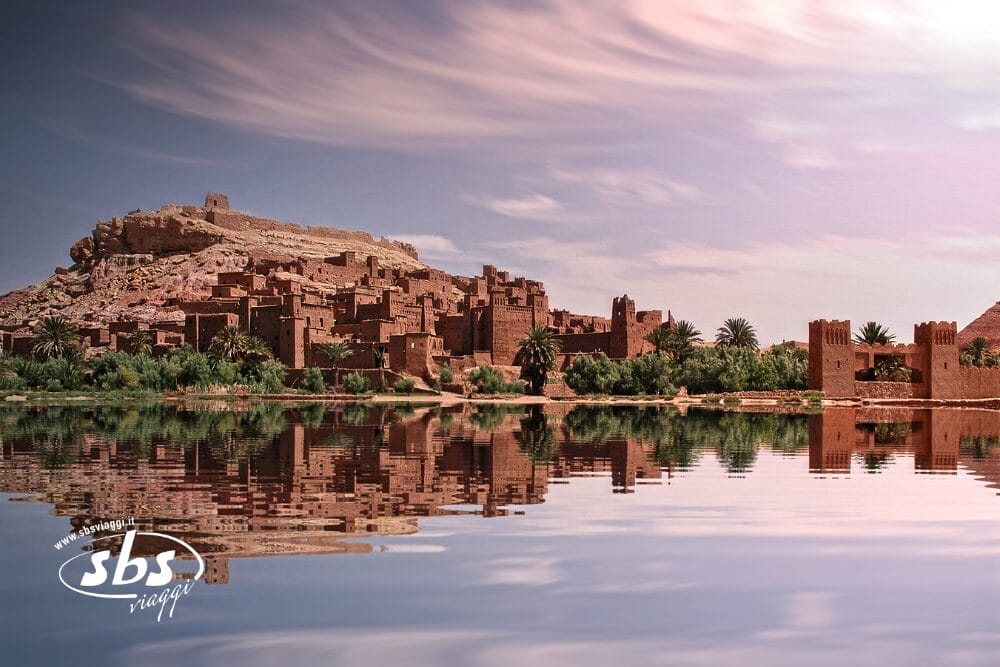 Veduta di un'antica città d'argilla con edifici marroni e mura fortificate, riflessa in un'acqua calma sotto nuvole soffici. Scoprite il meglio del Marocco in tour - il logo "sbs viaggi" appare nell'angolo in basso a sinistra.