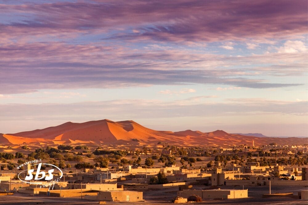 Una città con edifici dai tetti piatti si trova in primo piano, mentre dune di sabbia dorata e vegetazione sparsa si estendono oltre, catturando lo spirito di un Viaggio Marocco al tramonto sotto un cielo drammatico e parzialmente nuvoloso.