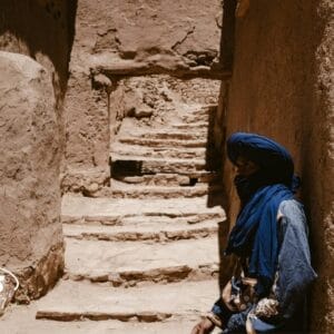 Una persona con turbante e sciarpa blu siede all'ombra di un vicolo stretto e illuminato dal sole, con gradini di pietra grezza e muri di terra, catturando l'essenza di un tour in Marocco. Il logo SBS è visibile nell'angolo.