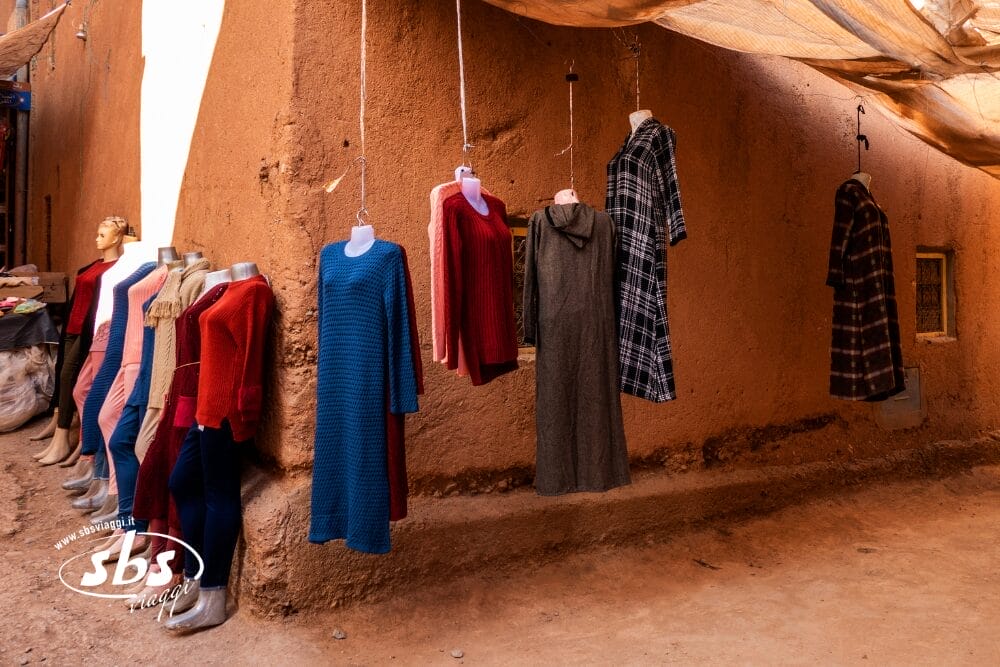 Manichini espongono abiti e maglioni a maniche lunghe in rosso, blu, marrone, grigio e a quadri all'esterno di un muro di adobe in un mercato all'aperto in Marocco, con alcuni capi di Tesori del Marocco appesi sopra.