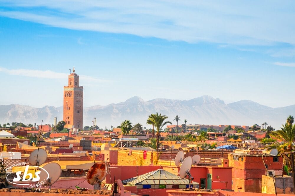 Veduta di Marrakech, in Marocco, con l'alto minareto della Moschea della Koutoubia, i tradizionali edifici rossastri con le antenne paraboliche, le palme e le montagne: una scena iconica di un'avventura di Tour Marocco sotto un cielo azzurro e limpido.