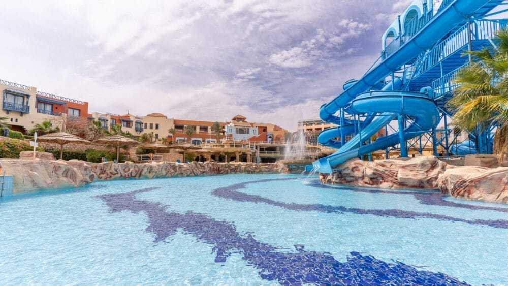 Una grande piscina all'aperto con uno scivolo blu e bordi rocciosi si trova a Faraana Heights, circondata da edifici colorati sotto un cielo parzialmente nuvoloso. Sullo sfondo sono visibili palme e lettini.