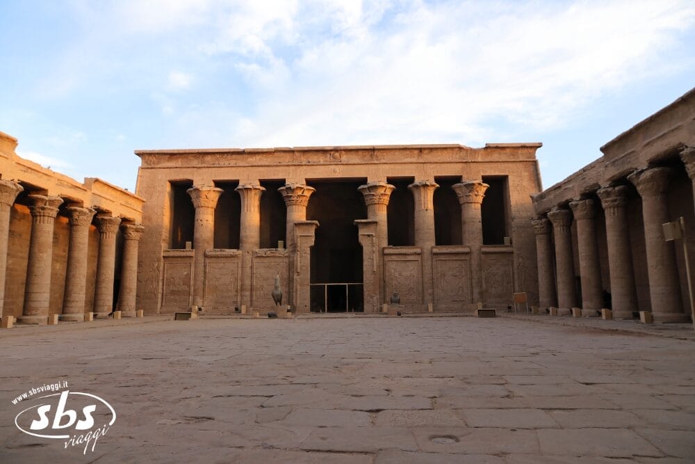 Cortile di un antico tempio egizio con alte colonne di pietra e sculture dettagliate, immerso in una calda luce solare, con cielo aperto e ombre sul terreno. Il logo "sbs viaggi" e Crociere Sul Nilo sono visibili nell'angolo in basso a sinistra.