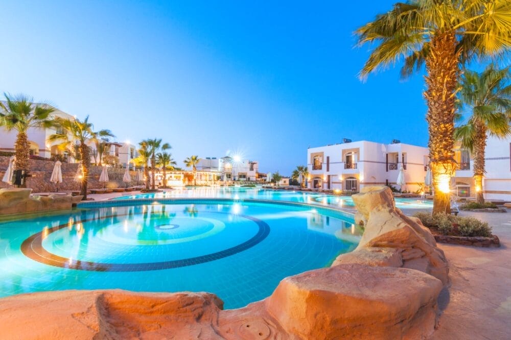 La lussuosa piscina dell'Amphoras Blu Resort è circondata da palme ed edifici bianchi e moderni al tramonto, con acqua cristallina, rocce e sedie a sdraio. Luci calde illuminano l'atmosfera rilassante.