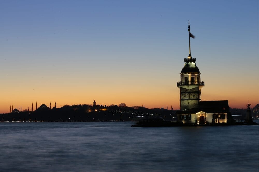 Una vista della Torre della Fanciulla illuminata al tramonto, in piedi sull'acqua con lo skyline della città di Istanbul e le moschee che si stagliano sotto un tramonto colorato: una scena iconica da vivere in un Tour Della Turchia.