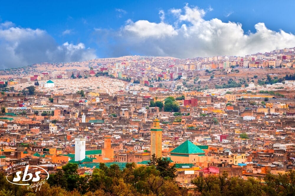 Una vista panoramica di Fez, in Marocco, mostra i tradizionali edifici abbronzati con i tetti verdi e le moschee - una scena invitante per un Tour Del Marocco - mentre le case colorate sulle colline riposano sotto un cielo blu parzialmente nuvoloso.