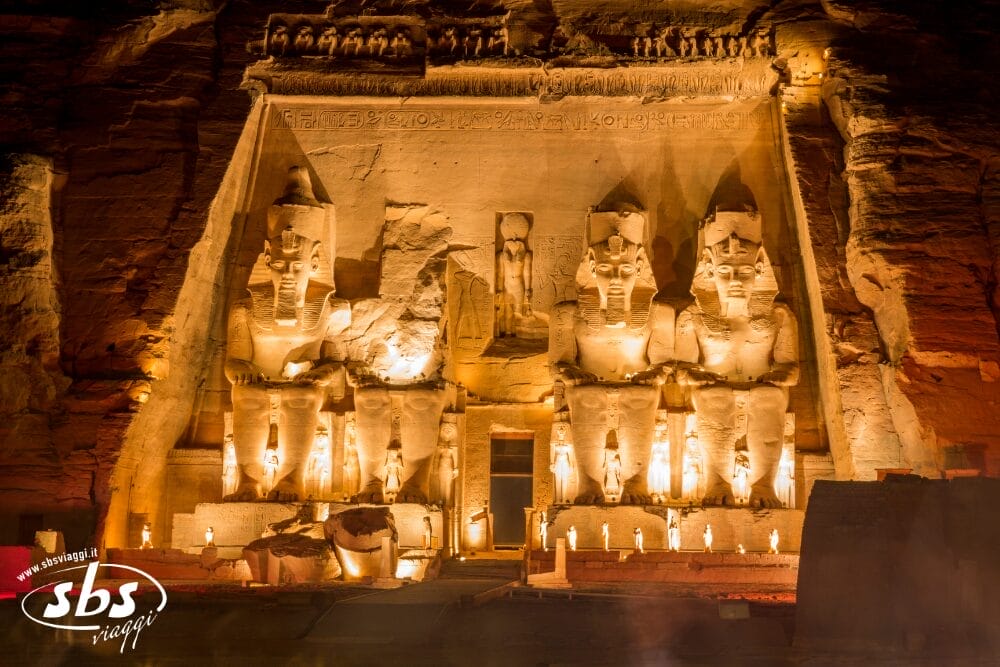Vista notturna del tempio illuminato di Abu Simbel in Egitto, un punto di forza di Crociere Sul Nilo, con quattro grandi statue sedute di Ramses II scolpite nella roccia, statue più piccole e geroglifici in alto.