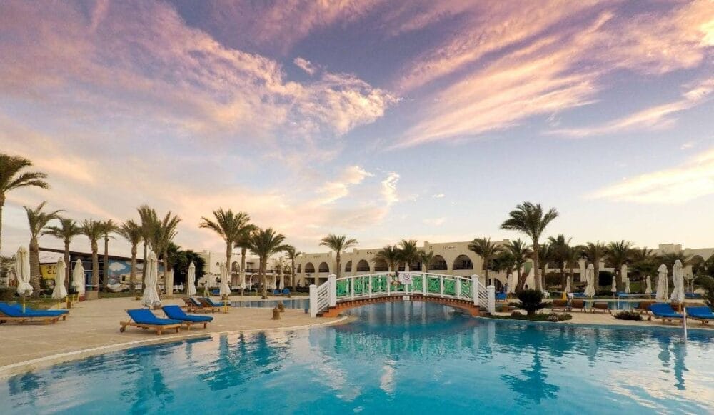 Una piscina del resort con sedie a sdraio blu, palme e una passerella bianca sotto il cielo colorato del tramonto all'Hilton Nubian Village. Nuvole rosa e arancioni illuminano la scena, con eleganti edifici bianchi sullo sfondo.