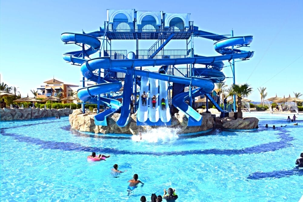 Una grande struttura acquatica blu a Faraana Heights presenta scivoli multipli e tortuosi, con i bambini che sguazzano in una piscina all'aperto di colore blu chiaro circondata da bagnanti e palme sullo sfondo.