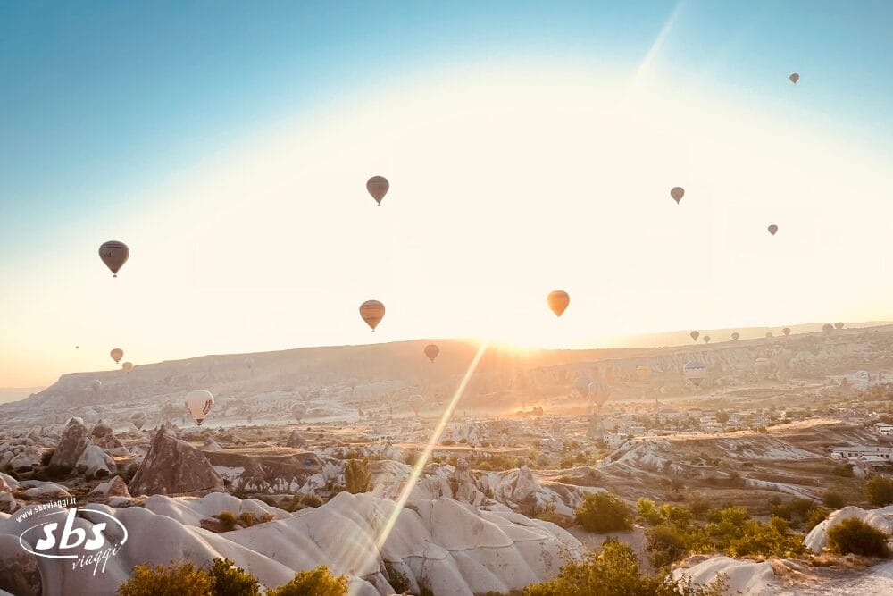 Diverse mongolfiere fluttuano in un cielo limpido all'alba sul paesaggio roccioso della Cappadocia, con una vegetazione sparsa in basso: una scena mozzafiato che spesso viene vissuta durante un Mini Tour Cappadocia.