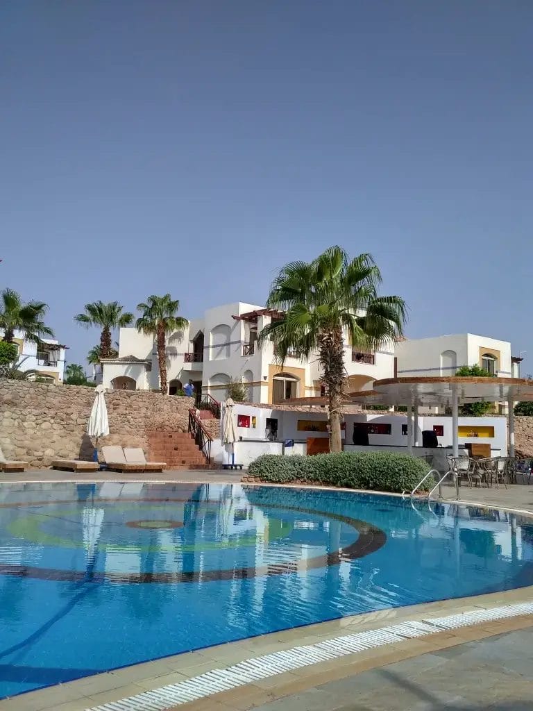 All'Amphoras Blu Resort vi attende una limpida piscina all'aperto con pavimento a motivi, circondata da sedie a sdraio, palme e ville bianche in stile mediterraneo sotto un cielo azzurro e soleggiato.