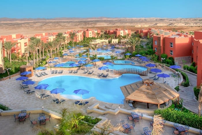 Un grande resort dispone di diverse piscine all'aperto con ombrelloni blu, sedie a sdraio, palme, edifici rossi e un ponte di legno, sullo sfondo delle colline del deserto sotto un cielo azzurro.