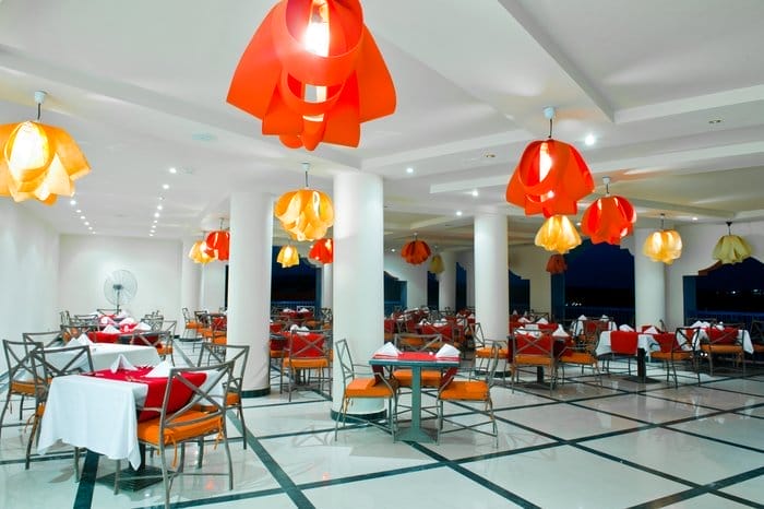 Interno di un ristorante moderno con pilastri bianchi, tavoli ricoperti di stoffe bianche e tovaglioli rossi, sedie in metallo con cuscini arancioni e grandi luci a sospensione arancioni e gialle appese al soffitto.