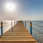 Il pontile in legno con pali blu e gialli dell'Aurora Oriental Resort si estende su acque calme e limpide verso il sole splendente all'orizzonte, sotto un cielo azzurro e terso.