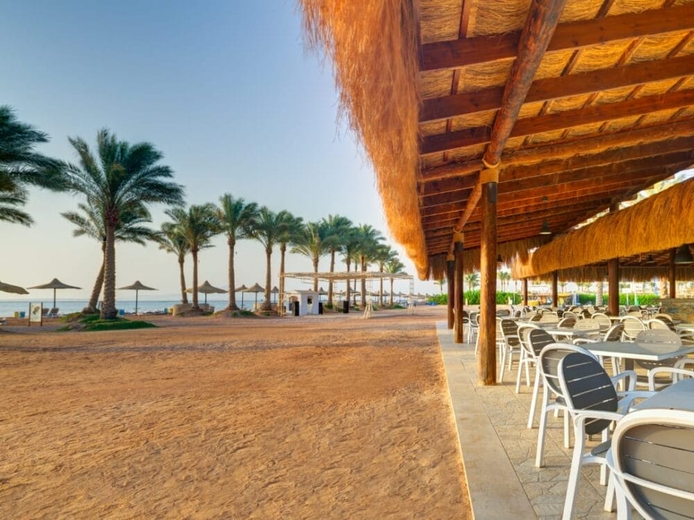 Un caffè all'aperto in riva al mare con tavoli e sedie bianche sotto un tetto di paglia, che si affaccia su una spiaggia sabbiosa fiancheggiata da palme e ombrelloni di paglia vicino al mare calmo dell'Aurora Oriental Resort di Sharm El Sheikh.