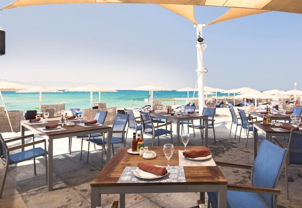 Ristorante all'aperto sul mare del Barcelo Tiran Sharm con tavoli in legno, sedie blu e ombrelloni. Godetevi una cena con vista sul mare turchese e ombrelloni sotto un cielo limpido in questo resort di lusso a Sharm El Sheikh.