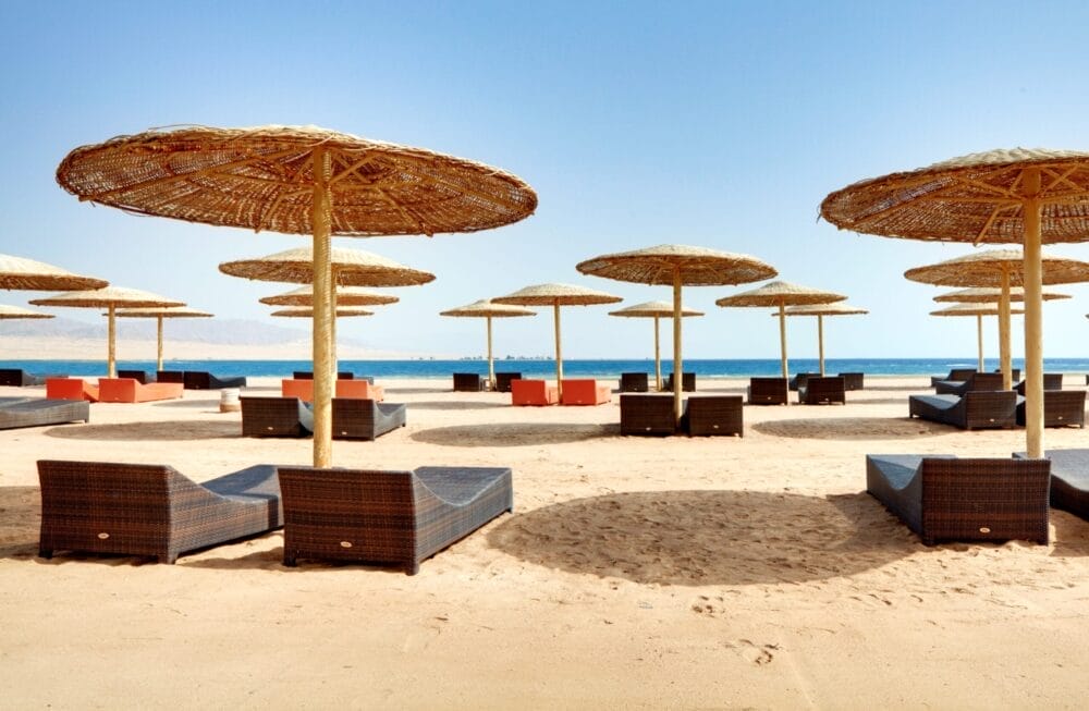 File di lettini di vimini e grandi ombrelloni di paglia sono disposti sulla spiaggia sabbiosa del Barcelo Tiran Sharm, di fronte al mare azzurro e calmo sotto un cielo limpido. La tranquilla spiaggia del resort Barcelo attende i visitatori.