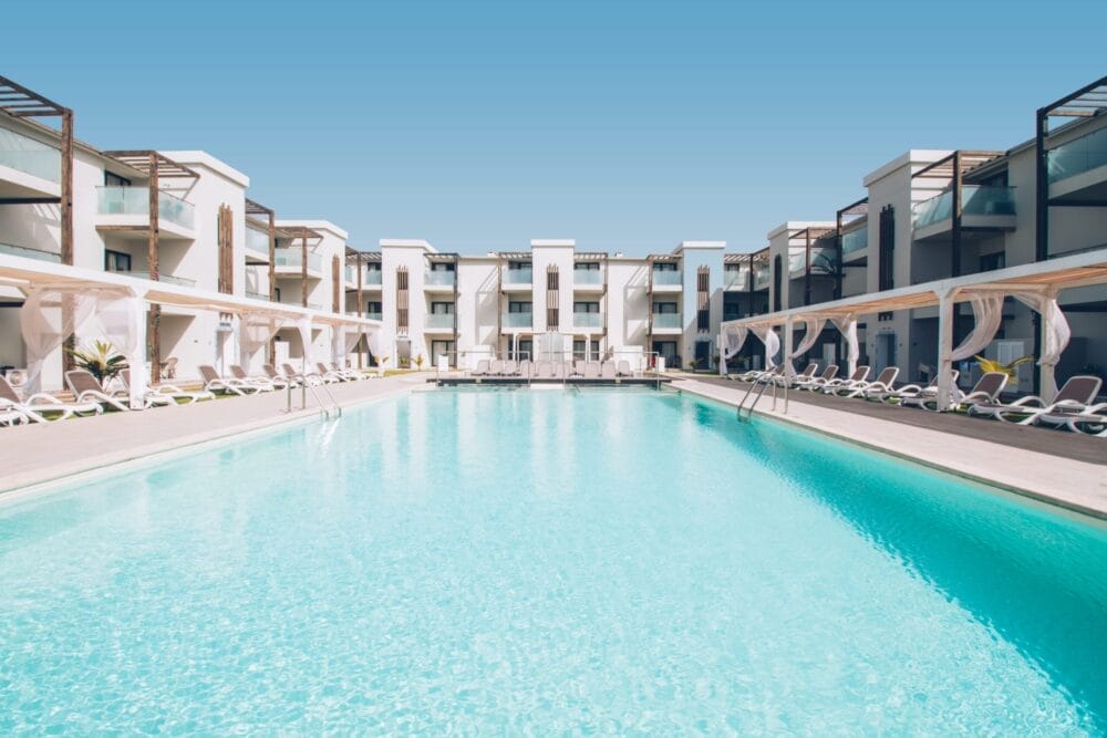 Una grande piscina trasparente è circondata da edifici moderni e bianchi con balconi e sedie a sdraio a bordo piscina sotto le tettoie dell'Halos Casa Resort, che si crogiolano sotto un cielo blu brillante.