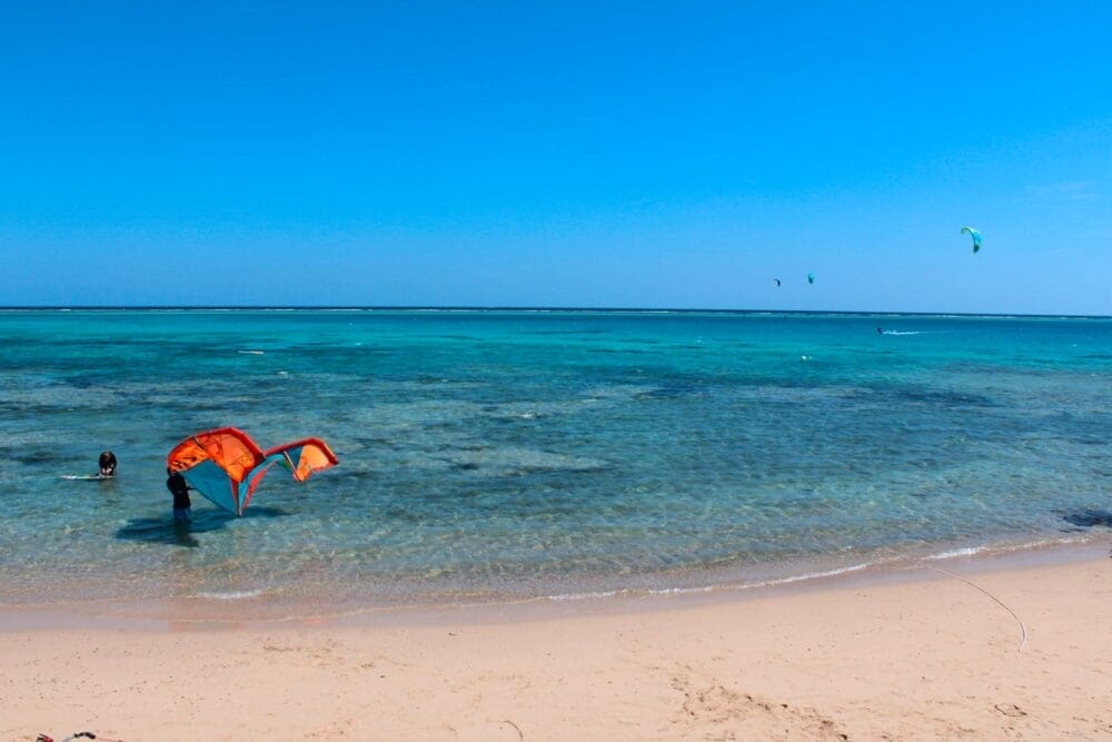 Una persona si trova nell'acqua azzurra e poco profonda del Dream Lagoon Resort e tiene in mano un aquilone arancione su una spiaggia sabbiosa, con tre kitesurfisti visibili in lontananza sotto un cielo azzurro brillante.