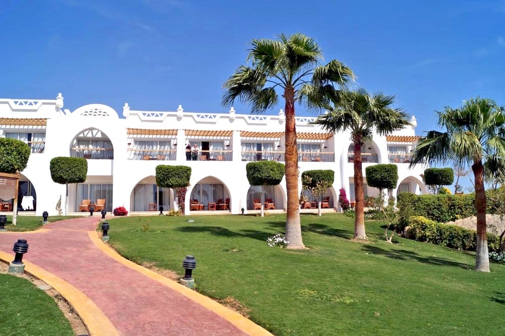 L'edificio bianco a due piani con balconi ad arco del Futura Club Albatros Palace, siepi curate, palme e una passerella rossa che si snoda attraverso un prato verde sotto un cielo azzurro e limpido.