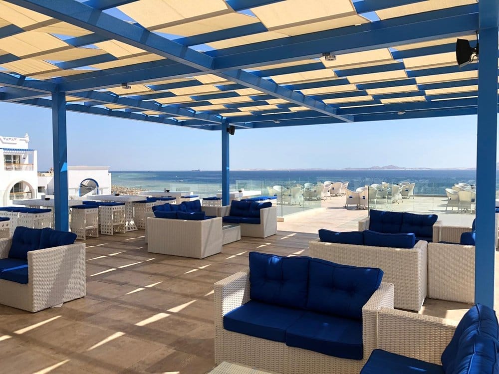 La terrazza sul mare del Futura Club Albatros Palace è caratterizzata da divani in vimini bianchi con cuscini blu sotto un pergolato blu, con vista sull'oceano calmo e limpido, con edifici bianchi e ringhiere di vetro sullo sfondo.