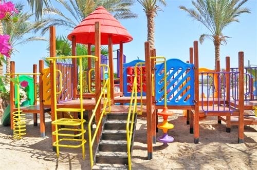 Una colorata struttura da gioco con scivoli, scale e arrampicate si trova al Futura Club Brayka Bay Resort, circondata da palme e fiori luminosi sotto un cielo azzurro e limpido. L'area giochi di sabbia è vuota.