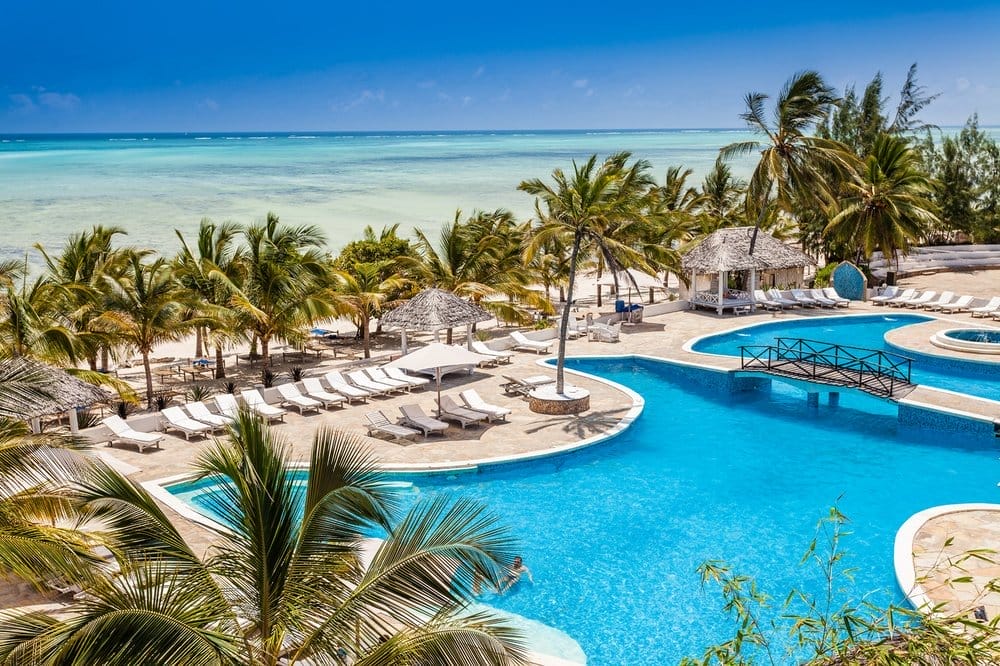 Vivete un'esperienza paradisiaca al Futura Club Twiga, un resort sulla spiaggia tropicale con una grande piscina blu, sedie a sdraio bianche, palme ondeggianti e una vista mozzafiato sull'oceano turchese sotto un cielo limpido. Un'incantevole passerella attraversa parte della piscina.