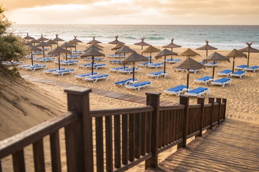 Scale di legno conducono alla spiaggia sabbiosa di Vila Baleira, fiancheggiata da file di sedie a sdraio blu e bianche e ombrelloni di paglia. L'oceano è calmo sotto un cielo arancione al tramonto.