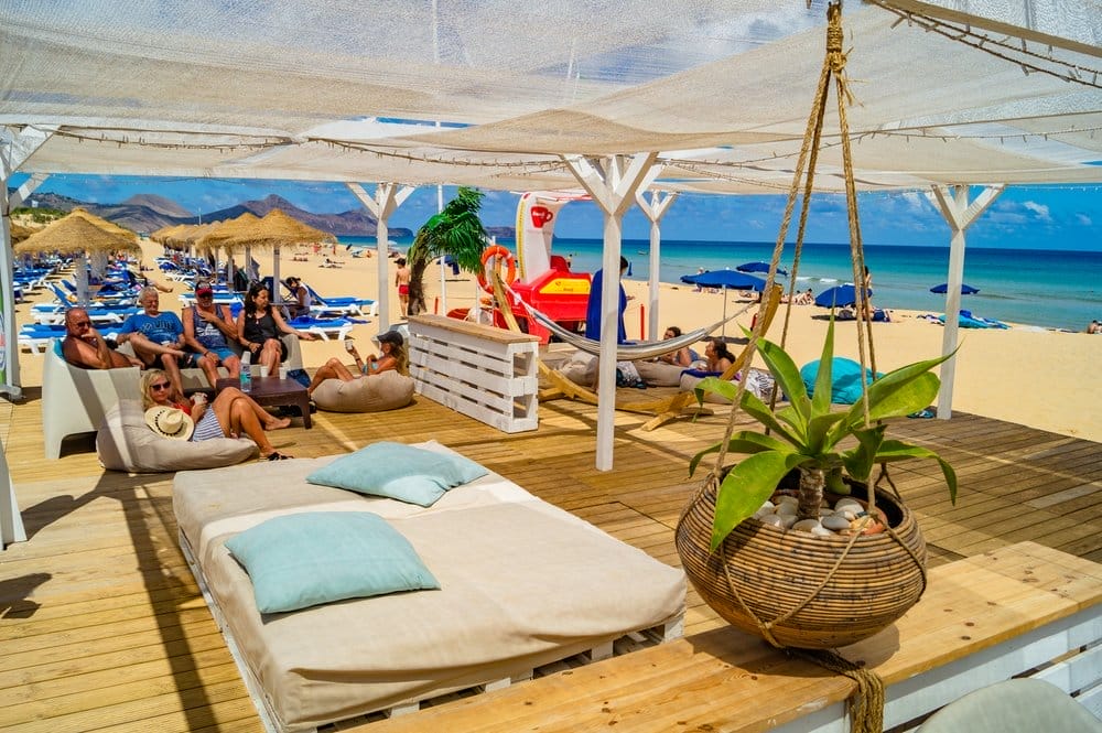 Le persone si rilassano su sedie a sdraio e sacchi di fagioli sotto una tettoia ombreggiata al Club Vila Baleira beach club. La spiaggia sabbiosa presenta ombrelloni blu, un'area giochi e viste sul mare, mentre una pianta pensile decora il primo piano.
