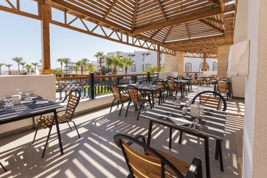 Patio del ristorante all'aperto del Nicolaus Club Remal Resort con tavoli neri e sedie in legno sotto un pergolato in legno che proietta ombre a strisce. Sullo sfondo, in una giornata di sole, si intravedono palme ed edifici bianchi.