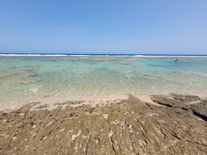 L'acqua turchese, limpida e poco profonda, incontra una costa rocciosa e sabbiosa sotto un cielo blu brillante; una persona nuota in lontananza vicino all'orizzonte del Three Corners Sea Beach Resort.
