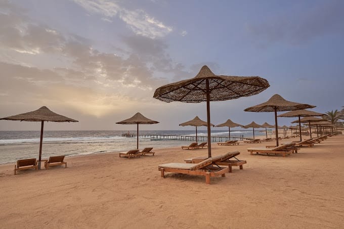 Sedie a sdraio in legno e ombrelloni di paglia sono disposti su una spiaggia sabbiosa al tramonto al Three Corners Sea Beach Resort, con onde calme e un molo sullo sfondo sotto un cielo parzialmente nuvoloso.