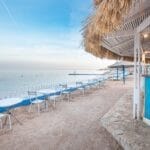 Il caffè sulla spiaggia dell'hotel Pyramisa è caratterizzato da tavoli e sedie bianche accanto all'acqua blu e calma. Ombrelloni di paglia offrono ombra, con un bar in legno colorato in primo piano. Il cielo limpido esalta la tranquilla atmosfera balneare.