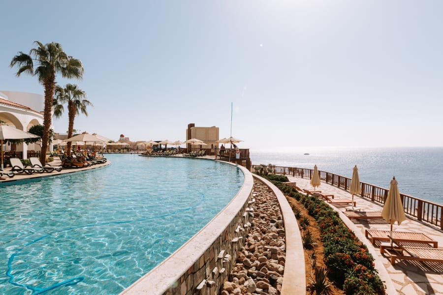 La grande piscina del resort Oasis Blue Bay si snoda tra lettini e ombrelloni, con palme e sentieri paesaggistici che si affacciano su una luminosa e soleggiata vista sull'oceano. Il cielo azzurro e le acque calme creano un'atmosfera rilassante.