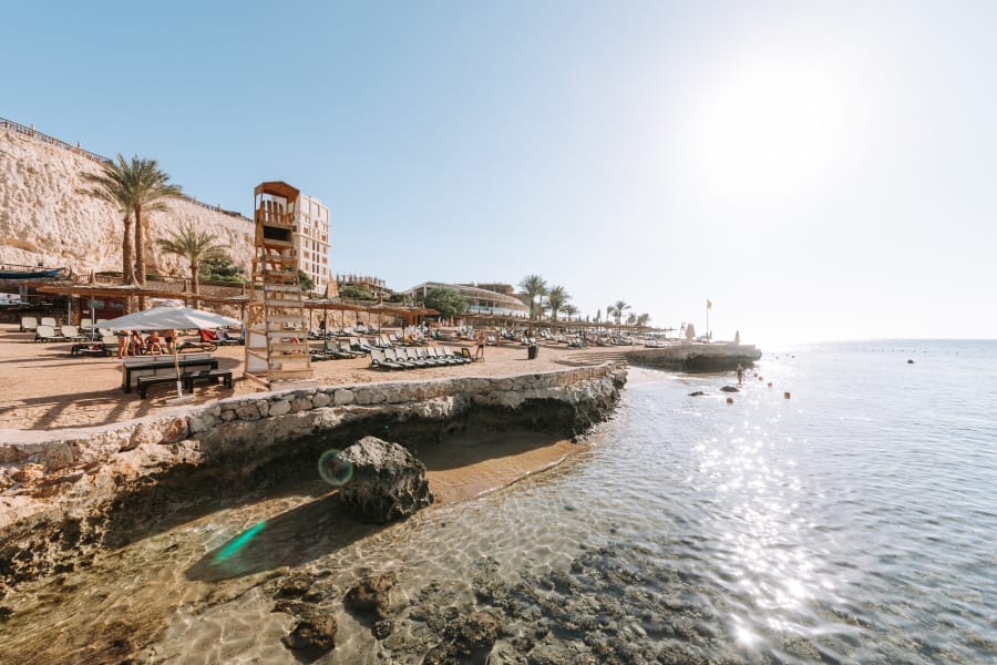 Una soleggiata località costiera di Sharm con acque azzurre e trasparenti, litorale roccioso, lettini, ombrelloni, una torre di salvataggio e palme. Alcune persone si rilassano sulla spiaggia di Blue Bay, incorniciata da edifici e scogliere sullo sfondo.